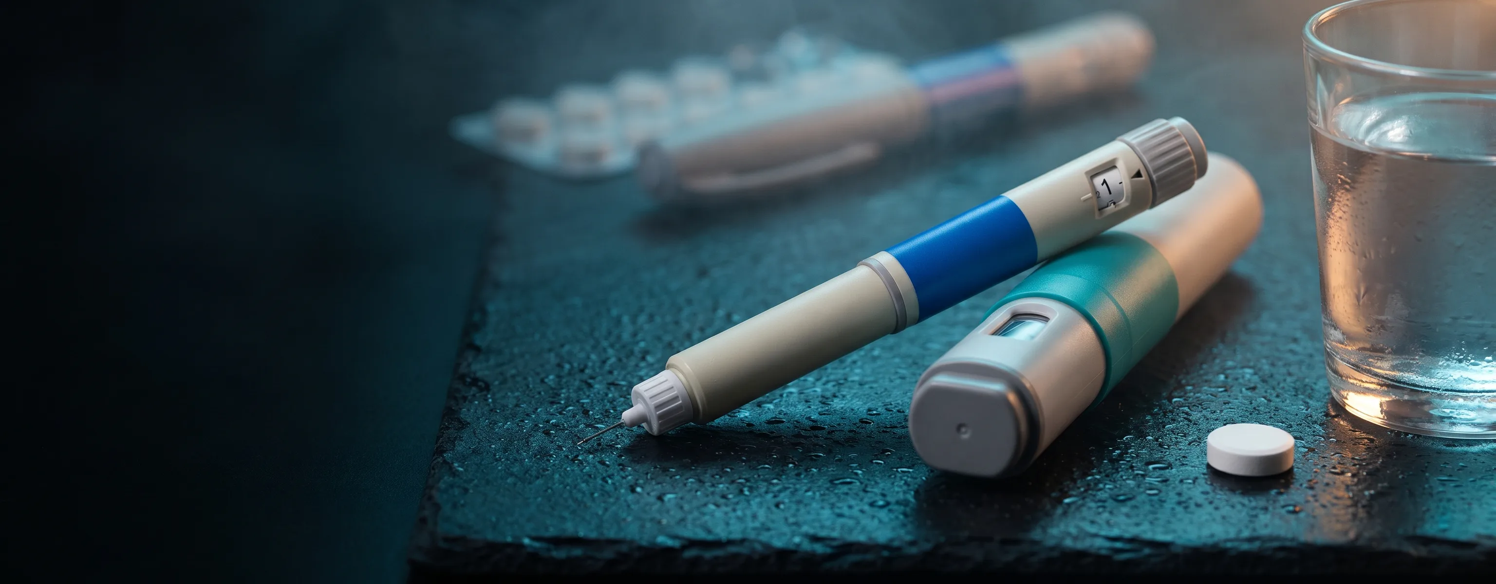 Two GLP-1 injection pens and a single tablet beside a glass of water on dark slate.